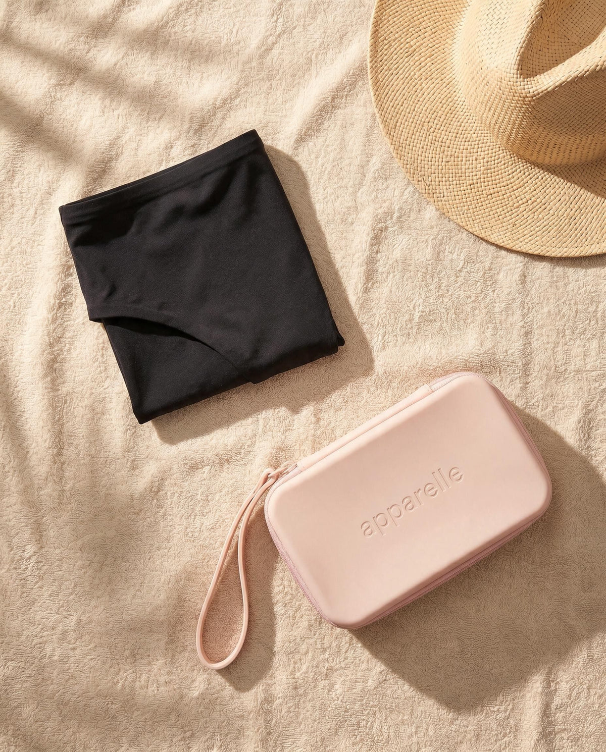 Braguita de bikini menstrual Apparelle doblada sobre una toalla junto al neceser impermeable rosa y un sombrero de paja