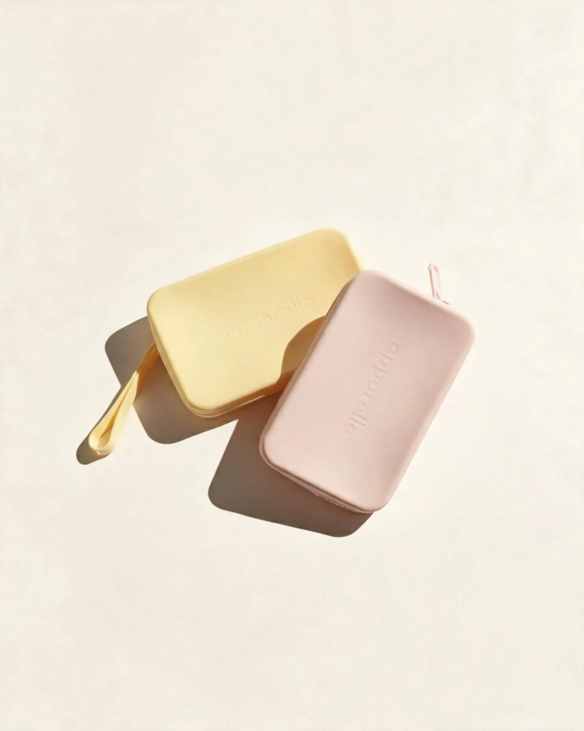 Dos neceseres impermeables de silicona Apparelle, en amarillo y rosa nude, con logotipo en relieve, sobre fondo crema